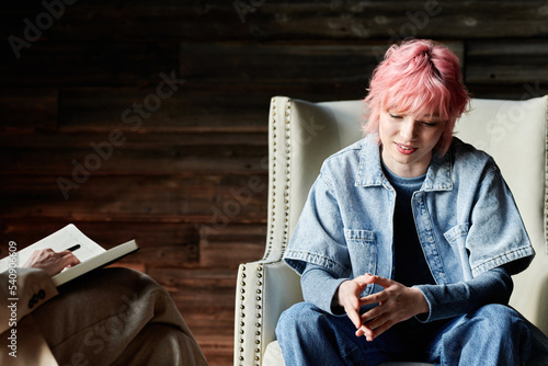 Teenage girl with pink hair sitting on chair at psychotherapy and talking to psychotherapist