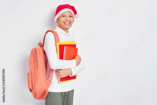 Cheerful handsome young Asian student in a Christmas hat wearing a t-shirt with backpack holding book isolated on white background