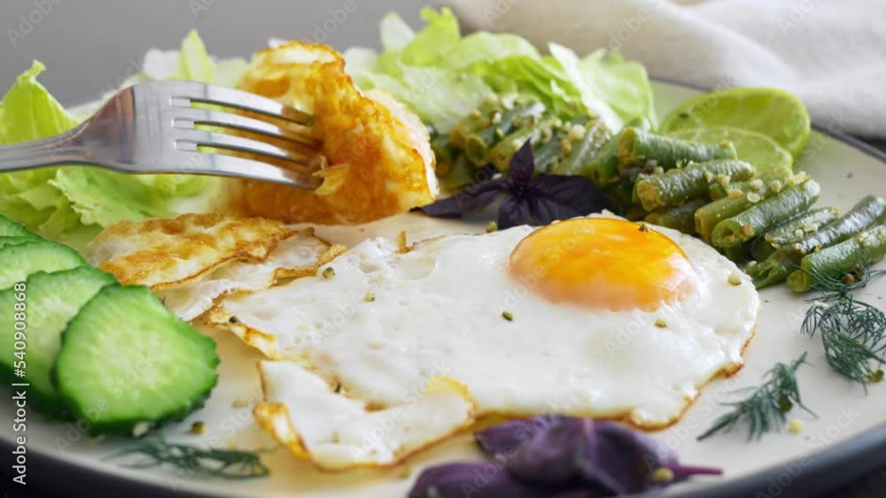 Close-up of a fork taking a fried egg yolk from a plate On a white plate, side view, a portion of fried eggs, fresh lettuce leaves, chopped cucumber and lemon slices, asparagus and greens Healthy food Close-up of a fork taking a fried egg yolk from a plate On a white plate, side view, a portion of fried eggs, fresh lettuce leaves, chopped cucumber and lemon slices, asparagus and greens Healthy food