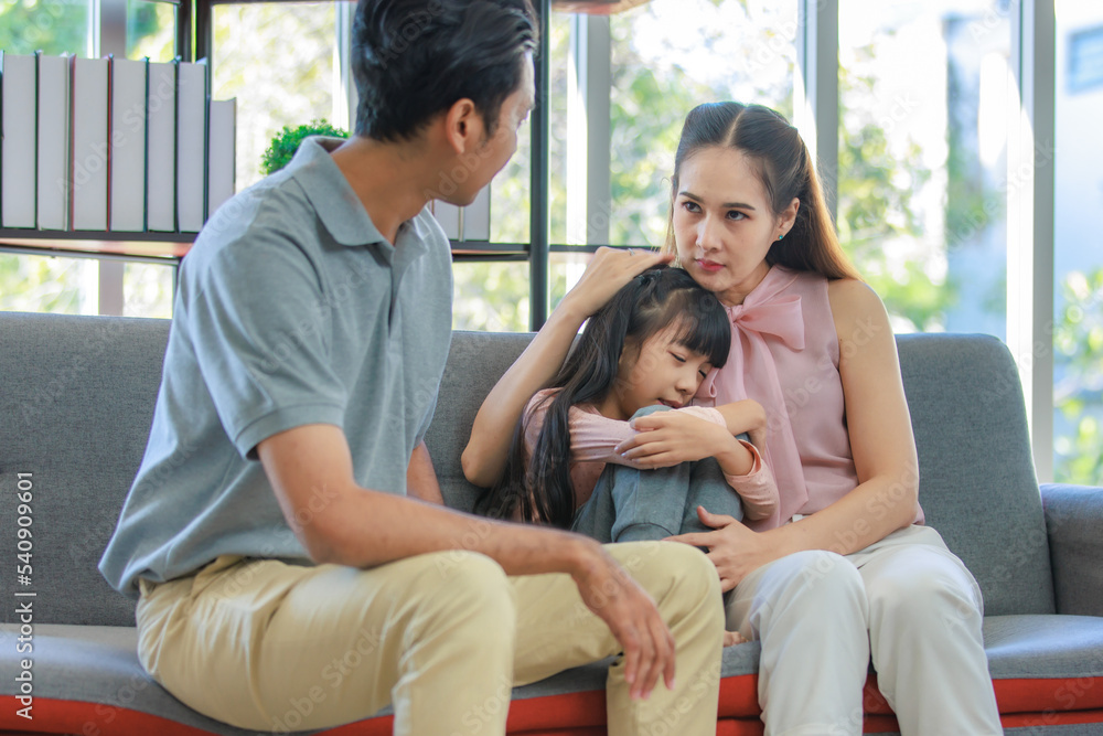 Millennial Asian unhappy family mother sitting on sofa couch holding hugging soothing little young female daughter while sad stressed father crying after big fight argument in living room at home Millennial Asian unhappy family mother sitting on sofa couch holding hugging soothing little young female daughter while sad stressed father crying after big fight argument in living room at home