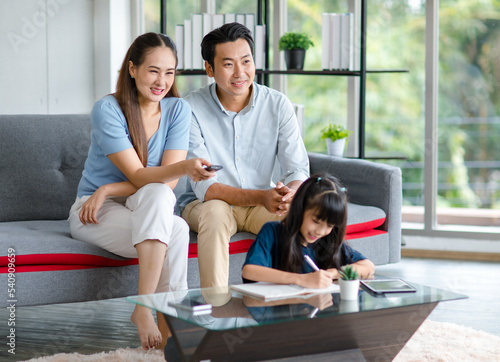 Millennial Asian happy family father and mother sitting on cozy sofa couch holding remote watching television while little young girl daughter sitting on floor doing homework in living room at home