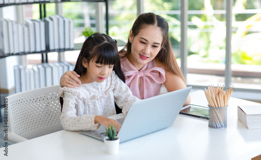 Fototapeta premium Millennial Asian happy family mother smiling helping supporting teaching little girl kid daughter studying learning doing online school homework via laptop notebook computer in living room at home