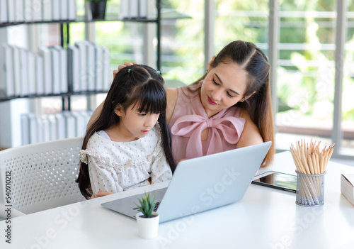 Millennial Asian happy family mother smiling helping supporting teaching little girl kid daughter studying learning doing online school homework via laptop notebook computer in living room at home