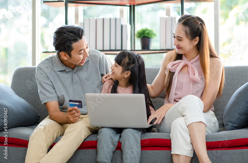 Asian happy excited little girl daughter sitting on cozy sofa couch smiling yelling screaming while father and mother using credit card making payment shopping gift present online via laptop computer