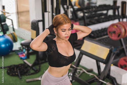 A young asian woman does prisoner squats at the gym. Hands on the back of the neck. Wearing a black crop top and fitness gloves. Training legs and lower body.