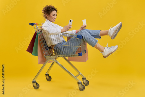 Side view of young woman smiling, taking selfie. Beautiful girl sitting in cart, holding packages, wearing white khudi, blue jeans and sneakers. Concept of carefree life.