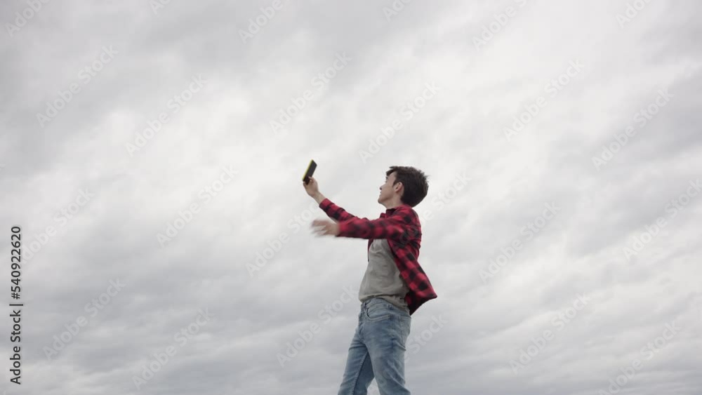 Influencer teenager holding phone and talking to camera outdoors. Boy streaming in a cloudy day. Wide angle view with copy space in the sky. Influencer teenager holding phone and talking to camera outdoors. Boy streaming in a cloudy day. Wide angle view with copy space in the sky.