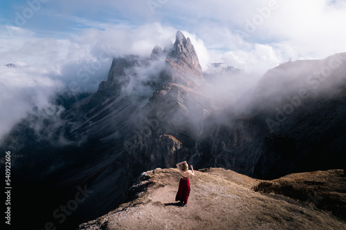A beautiful girl in a red dress stands facing the mountain partially covered with clouds under the blue sky. Autumn. Italian Alps, Dolomites.