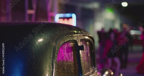 Beautiful old car roof and window covered in droplets of heavy rain. Blurry city lights , neon sign bokeh  background . Many people dancing in the rain. Colorful outdoor party at raining day . Closeup