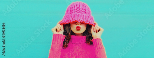 Winter colorful portrait of beautiful young woman blowing her lips wearing knitted sweater, pink hat on blue background