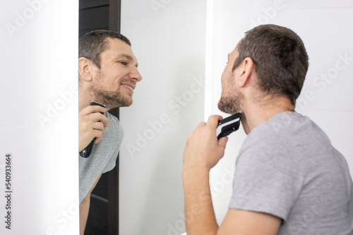 A man shaves his beard with a trimmer , looks in the mirror and smiles