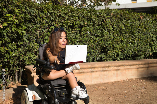 Disabled woman with reduced mobility and small stature in an electric wheelchair working with her laptop computer outdoors. Concept handicap, disability, incapacity, special needs, teleworking. photo