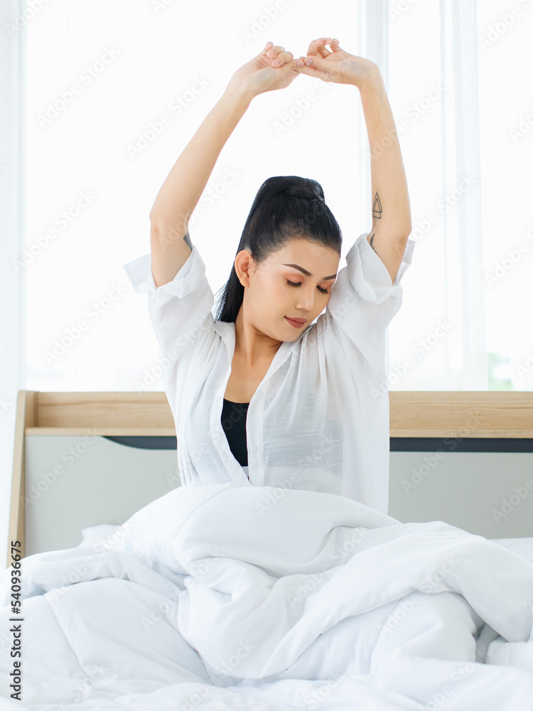 Portrait shot of Millennial Asian sexy female in casual comfort tanktop and shirt sitting under white thick warm blanket posing stretching arm after waking up in bedroom in morning at home. Portrait shot of Millennial Asian sexy female in casual comfort tanktop and shirt sitting under white thick warm blanket posing stretching arm after waking up in bedroom in morning at home.