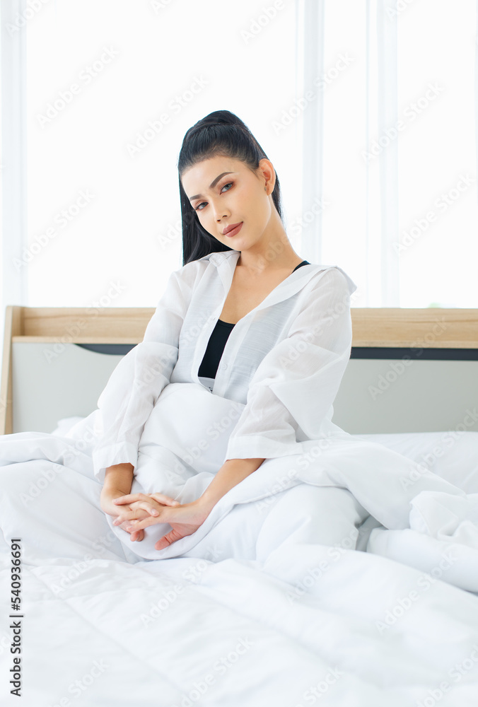 Portrait shot of Millennial Asian sexy female in casual comfort tanktop and shirt sitting under white thick warm blanket posing stretching arm after waking up in bedroom in morning at home. Portrait shot of Millennial Asian sexy female in casual comfort tanktop and shirt sitting under white thick warm blanket posing stretching arm after waking up in bedroom in morning at home.
