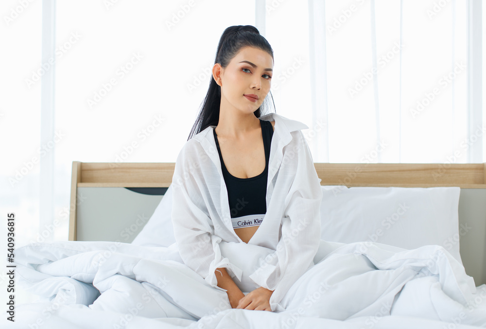 Portrait shot of Millennial Asian sexy female in casual comfort tanktop and shirt sitting under white thick warm blanket posing look at camera smiling after waking up in bedroom in morning at home Portrait shot of Millennial Asian sexy female in casual comfort tanktop and shirt sitting under white thick warm blanket posing look at camera smiling after waking up in bedroom in morning at home