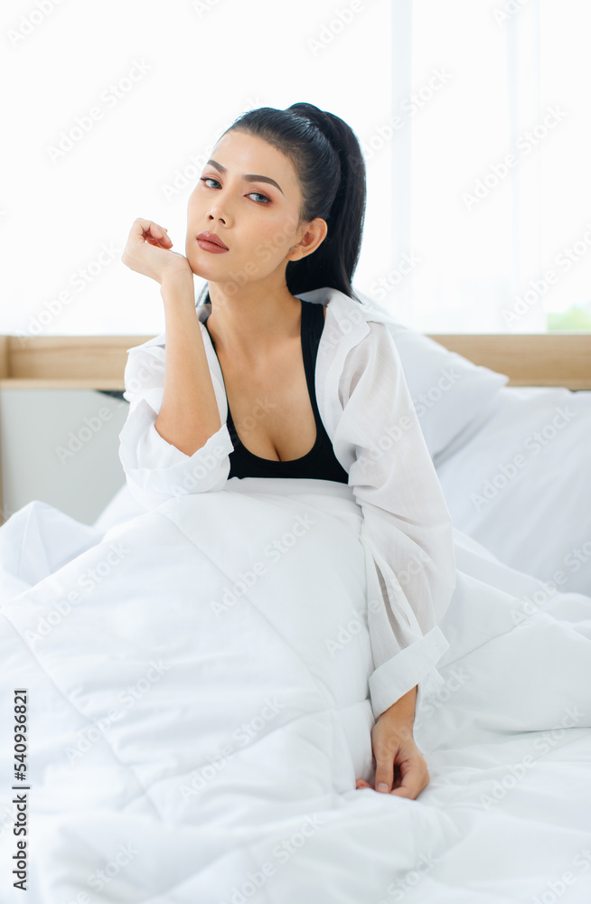 Portrait shot of Millennial Asian sexy female in casual comfort tanktop and shirt sitting under white thick warm blanket posing look at camera smiling after waking up in bedroom in morning at home Portrait shot of Millennial Asian sexy female in casual comfort tanktop and shirt sitting under white thick warm blanket posing look at camera smiling after waking up in bedroom in morning at home