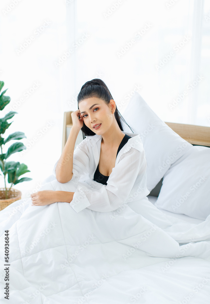 Portrait shot of Millennial Asian sexy female in casual comfort tanktop and shirt sitting under white thick warm blanket posing look at camera smiling after waking up in bedroom in morning at home Portrait shot of Millennial Asian sexy female in casual comfort tanktop and shirt sitting under white thick warm blanket posing look at camera smiling after waking up in bedroom in morning at home