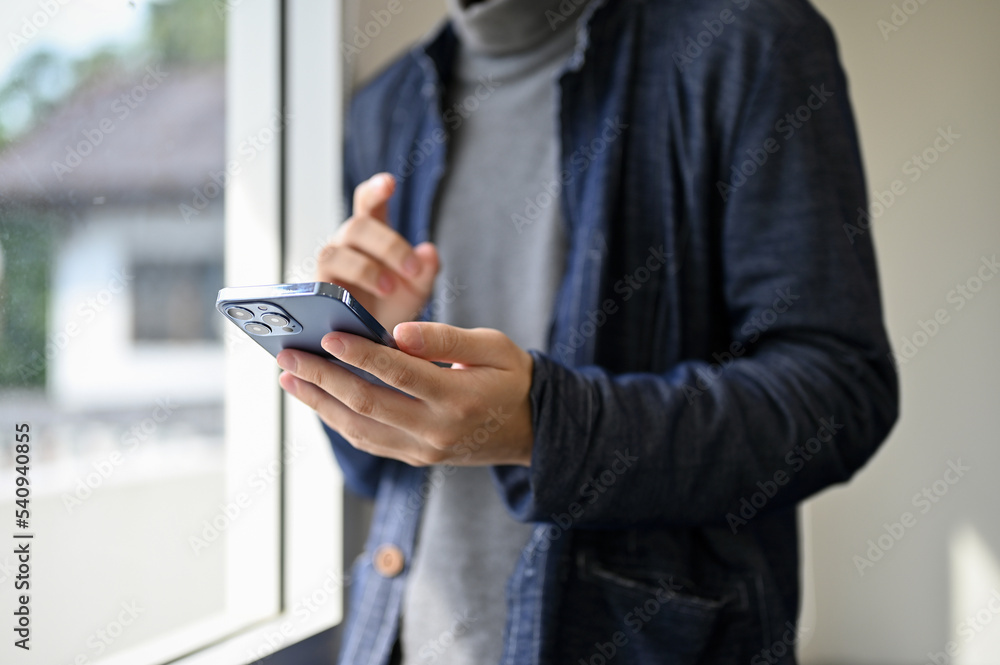 cropped, Asian man in casual clothes stands by the window, using his smart mobile phone