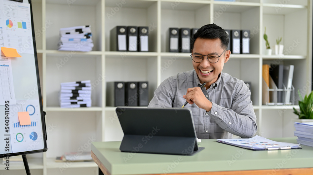 Cheerful millennial Asian businessman looking at his tablet screen, working at his desk. Cheerful millennial Asian businessman looking at his tablet screen, working at his desk.