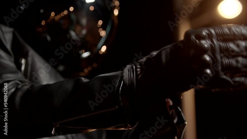 Biker In Leather Jacket, wearing , tying motorcycle black gloves and helmet . Biker Man Taking Off or installing protective clothes . Close up of rider sitting on beautiful motorcycle at night
