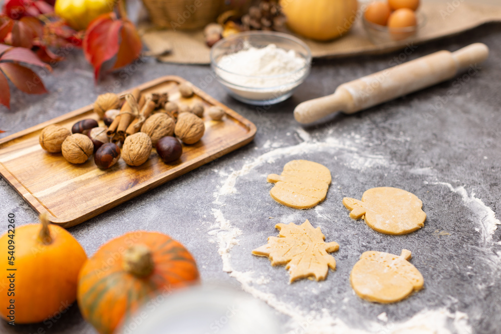 Cookies in the form of autumn leaves are rolled out of dough on the table. Nuts, leaves and pumpkins lie on the background. Thanksgiving cookies. Festive pastries for adults and children.