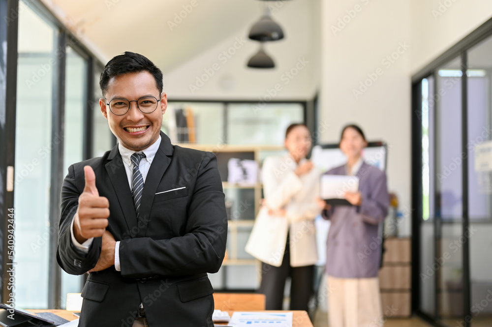 Professional adult Asian male boss showing thumb up, standing in his office building Professional adult Asian male boss showing thumb up, standing in his office building
