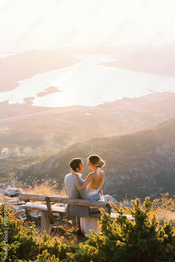 Bride sits on the lap of groom on a bench on top of a mountain