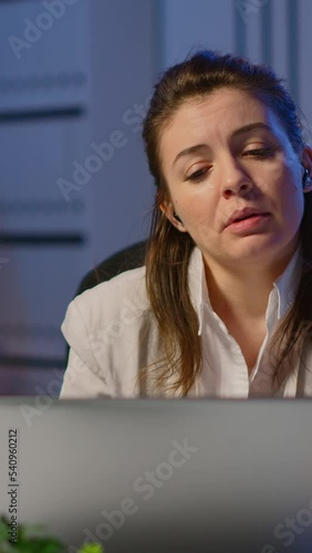 Vertical video: Business woman using wireless headphone duing video conference working overtime from start-up office in front of laptop. Lady using modern technology network talking on virtual meeting photo