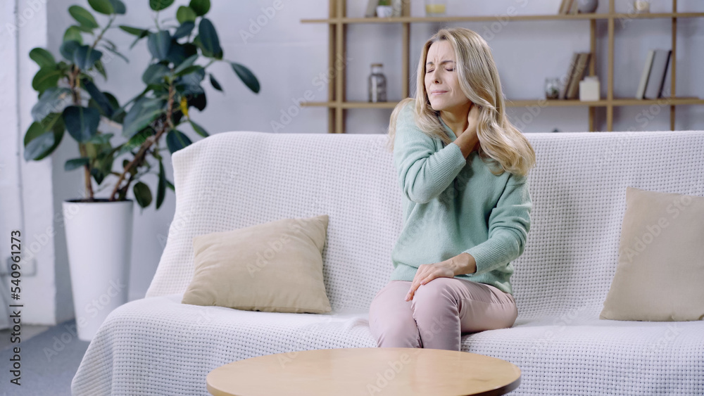 upset woman with closed eyes having pain in neck while sitting on sofa in modern living room upset woman with closed eyes having pain in neck while sitting on sofa in modern living room