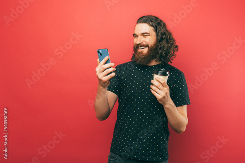 Photo of happy young handsome bearde man with long hair using smartphone and holding cup of coffee to go over pink background.