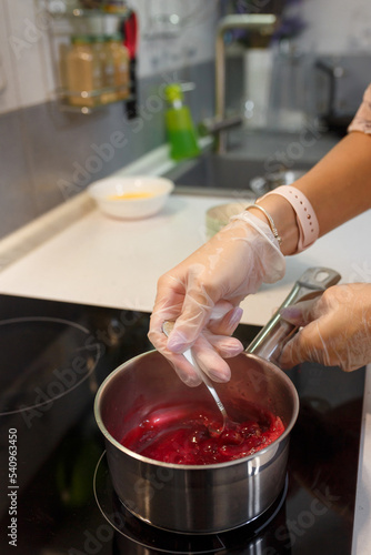 Making jam. Hands in gloves mixing sugar and berries with spoon in saucepot on stove. Seasonal berry jelly or marmalade. Kitchen background