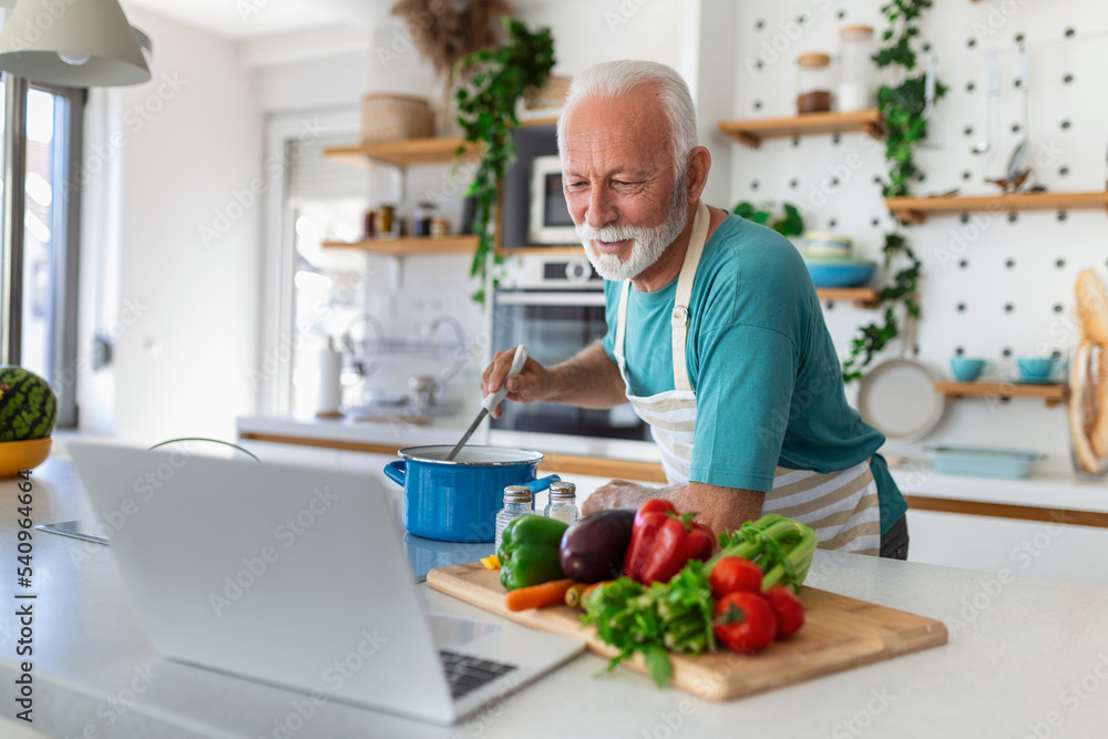 Happy senior man having fun cooking at home - Elderly person preparing healthy lunch in modern kitchen looking at the receipt at his laptop Happy senior man having fun cooking at home - Elderly person preparing healthy lunch in modern kitchen looking at the receipt at his laptop