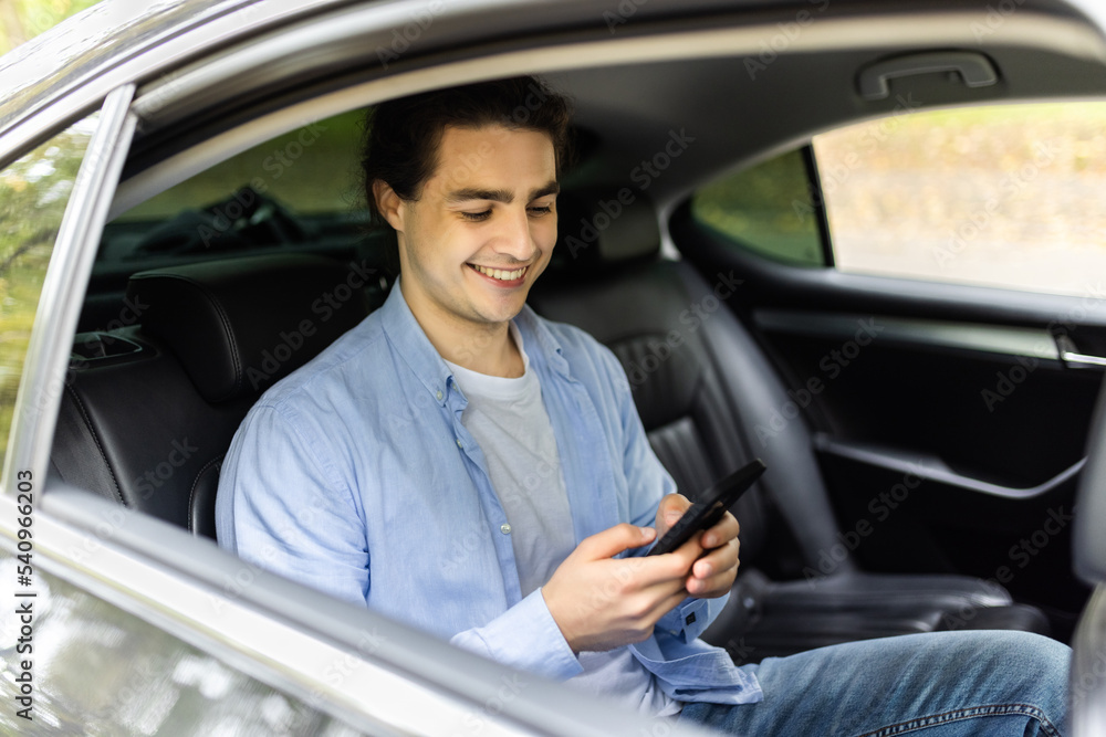 Happy smiling business man typing message on phone while sitting in a taxi. Young businessman clothing using smartphone while sitting on back seat in car. Cheerful guy messaging with cell.