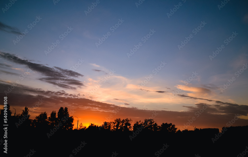 The morning glow illuminates the clouds on a beautiful colorful sky above the silhouettes of trees and a pillar at dawn