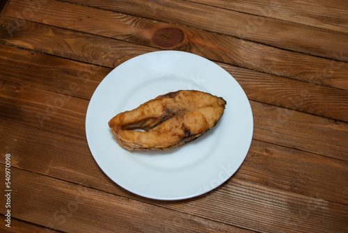A piece of fried river fish on a white plate that stands on a natural wood table