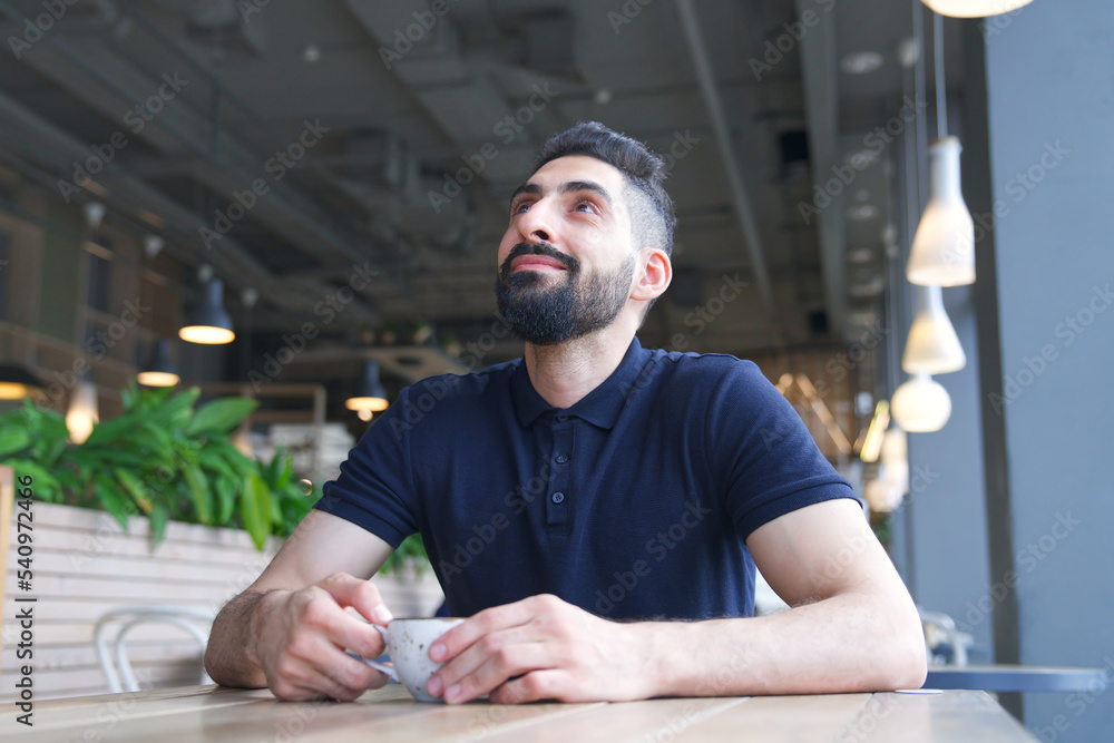 Portrait of happy positive Arab Muslim ethnic guy in glasses, young bearded man with black beard is drinking coffee sitting at table in cafe or restaurant, smiling Portrait of happy positive Arab Muslim ethnic guy in glasses, young bearded man with black beard is drinking coffee sitting at table in cafe or restaurant, smiling