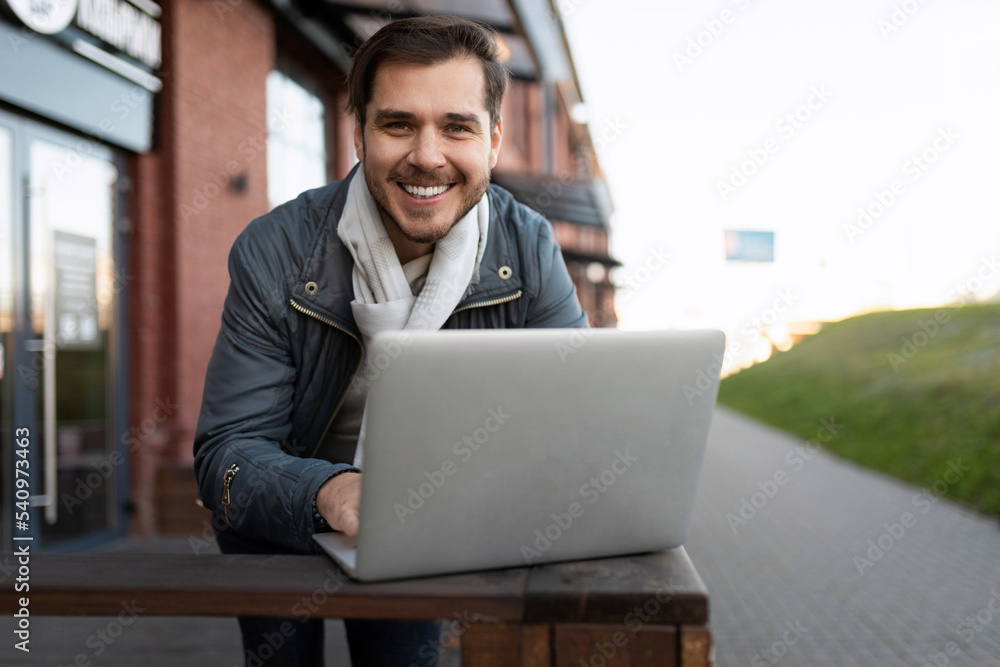a man with a laptop smiling broadly looks at the camera against the backdrop of a city building