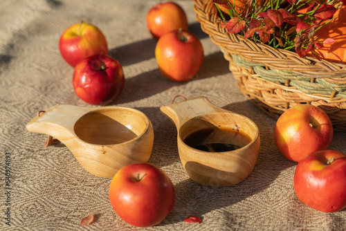 A variety of pumpkins in a wicker basket, red apples and tea in wooden mugs on a table covered with a canvas tablecloth. Autumn still life. Horizontal photo.