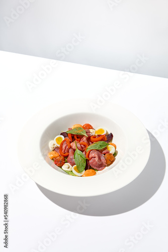 Salad with roast beef, vegetables and eggs on white table with harsh shadows. Steak salad in modern style with shadows of leaves. Flashy food. Meat salad with pastrami in summer menu.