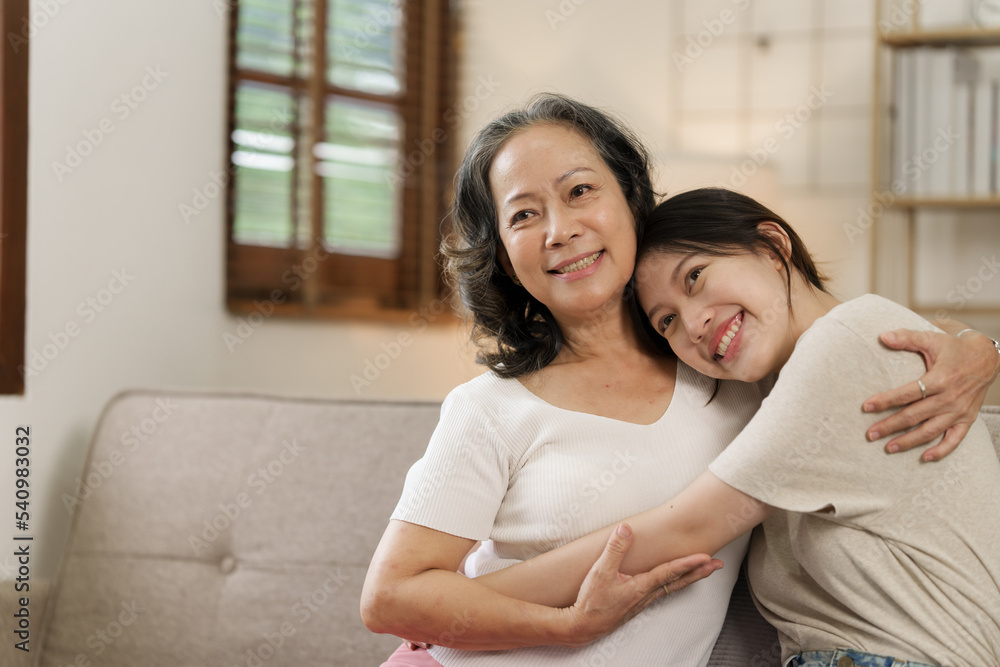 Elderly mother and grown daughter holding hands sitting on the sofa hugging.