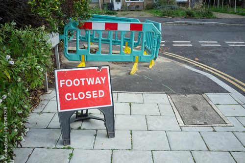 A red sign and barriers saying that footway ahead is closed as there are roadworks on the pavement. London, UK