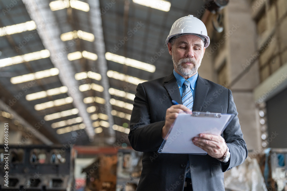 Senior businessman owner wearing helmet safety standing and writing on clipboard in heavy metal factory industry. Confident entrepreneur in suit checking lists on document at factory warehouse. Senior businessman owner wearing helmet safety standing and writing on clipboard in heavy metal factory industry. Confident entrepreneur in suit checking lists on document at factory warehouse.