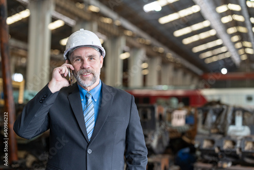 Smiling senior businessman wearing helmet safety standing and using smartphone in heavy metal factory industrial. Confident entrepreneur in suit talking with business partner. Work success concept.