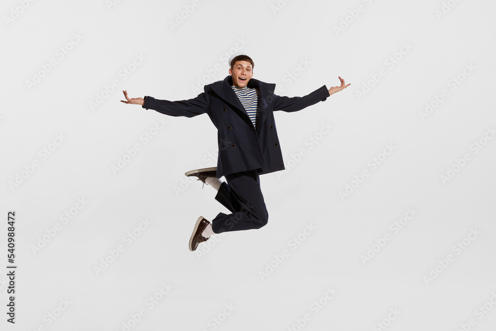 Portrait of young boy, sailor in striped shirt and jacket posing, jumping isolated over white background