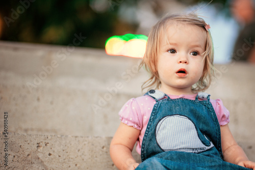 Street portrait of a little girl on a walk. Young blonde in the rays of the setting sun. A cute little girl is sitting on the steps of the pier on the seashore © watman