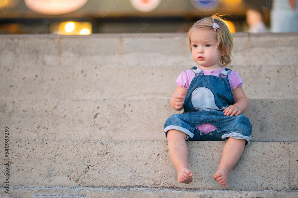 A cute little girl is sitting on the steps of the pier on the seashore. Young blonde in the rays of the setting sun. Street portrait of a little girl on a walk A cute little girl is sitting on the steps of the pier on the seashore. Young blonde in the rays of the setting sun. Street portrait of a little girl on a walk