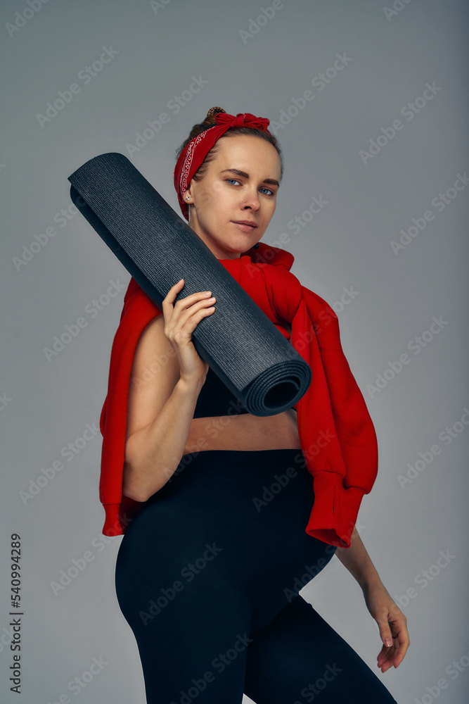 Pregnant woman holding yoga mat in hands, going for sport while awaiting baby, looking smiling at camera and touching her belly, standing against gray wall. Pregnant woman holding yoga mat in hands, going for sport while awaiting baby, looking smiling at camera and touching her belly, standing against gray wall.