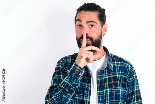 young bearded hispanic man wearing plaid shirt over white background makes hush gesture, asks be quiet. Don't tell my secret or not speak too loud, please!