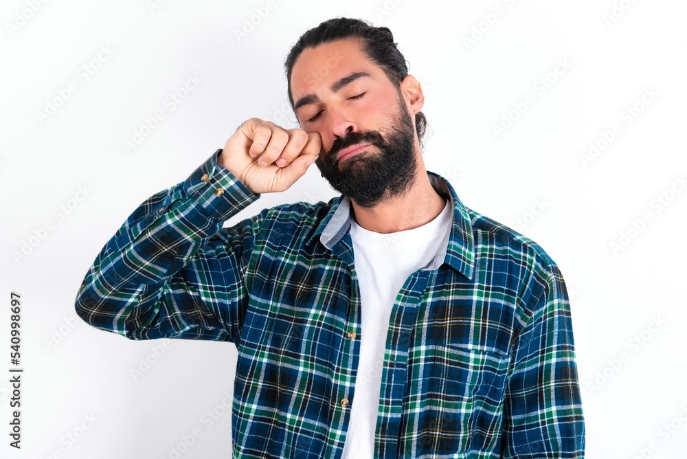 Unhappy young bearded hispanic man wearing plaid shirt over white background crying while posing at camera whipping tears with hand. Unhappy young bearded hispanic man wearing plaid shirt over white background crying while posing at camera whipping tears with hand.