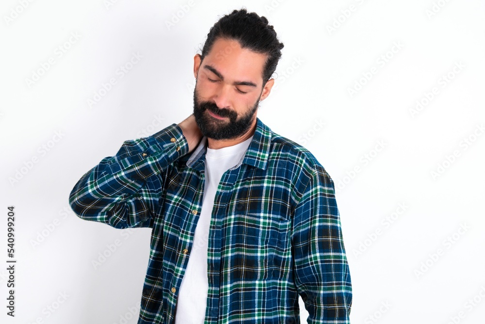 young bearded hispanic man wearing plaid shirt over white background suffering from back and neck ache injury, touching neck with hand, muscular pain. young bearded hispanic man wearing plaid shirt over white background suffering from back and neck ache injury, touching neck with hand, muscular pain.
